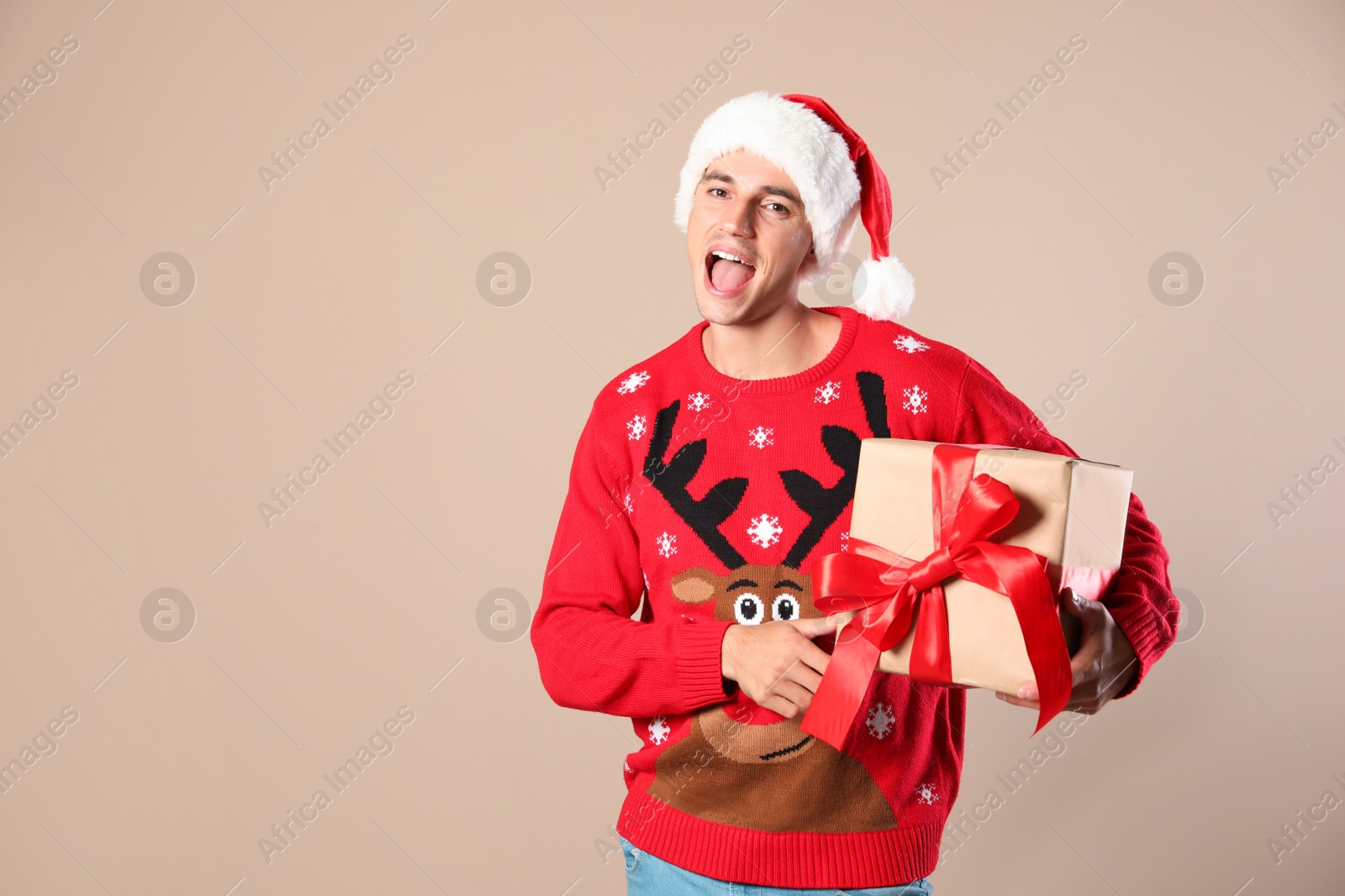 Photo of Happy man in Christmas sweater and Santa hat holding gift box on beige background