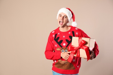 Photo of Happy man in Christmas sweater and Santa hat holding gift box on beige background