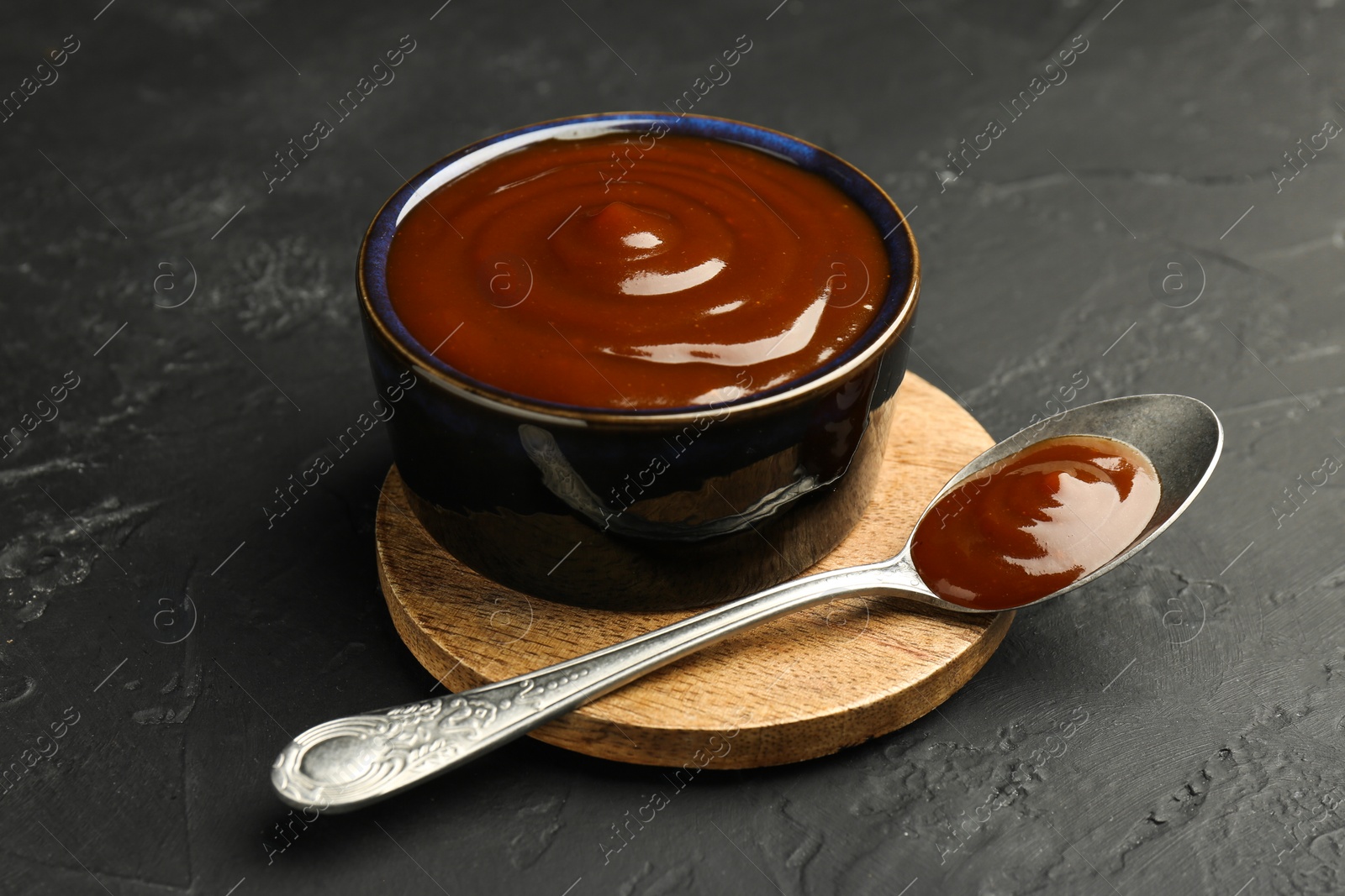 Photo of Tasty barbeque sauce in bowl and spoon on dark textured table, closeup