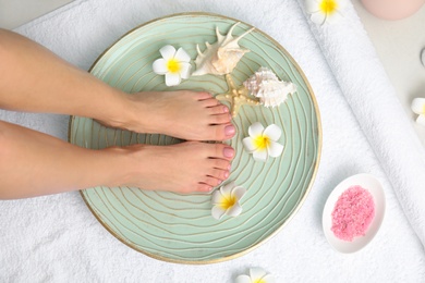 Photo of Woman soaking her feet in plate with water, flowers and seashells on white towel, top view. Spa treatment