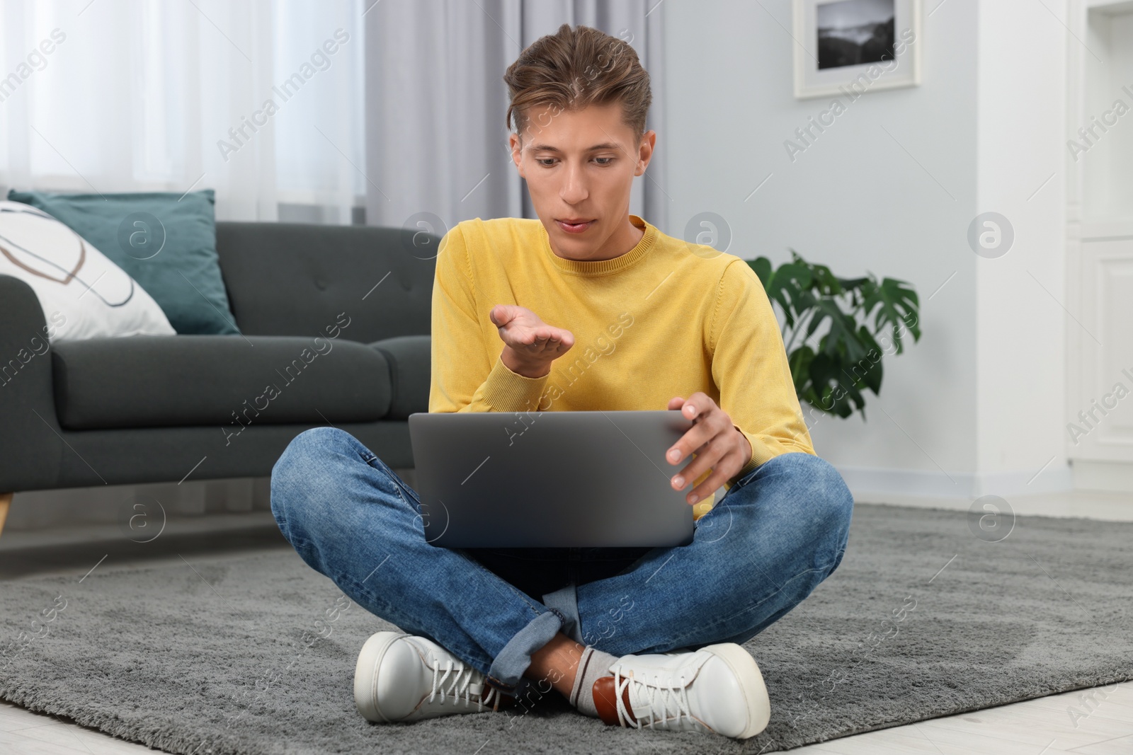 Photo of Happy young man having video chat via laptop and blowing kiss on carpet indoors. Long-distance relationship