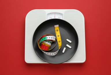 Photo of Scales with plate, weight loss pills and measuring tape on red background, top view