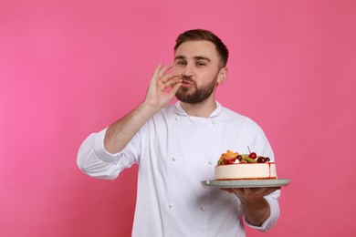 Happy professional confectioner in uniform with cake showing delicious gesture on pink background