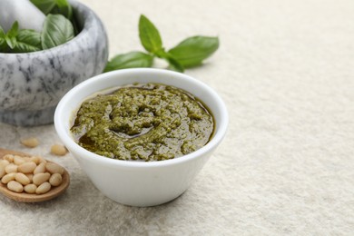 Photo of Tasty pesto sauce in bowl, basil, pine nuts and spoon on light grey table, space for text