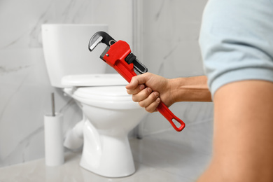 Photo of Professional plumber holding pipe wrench near toilet bowl in bathroom, closeup
