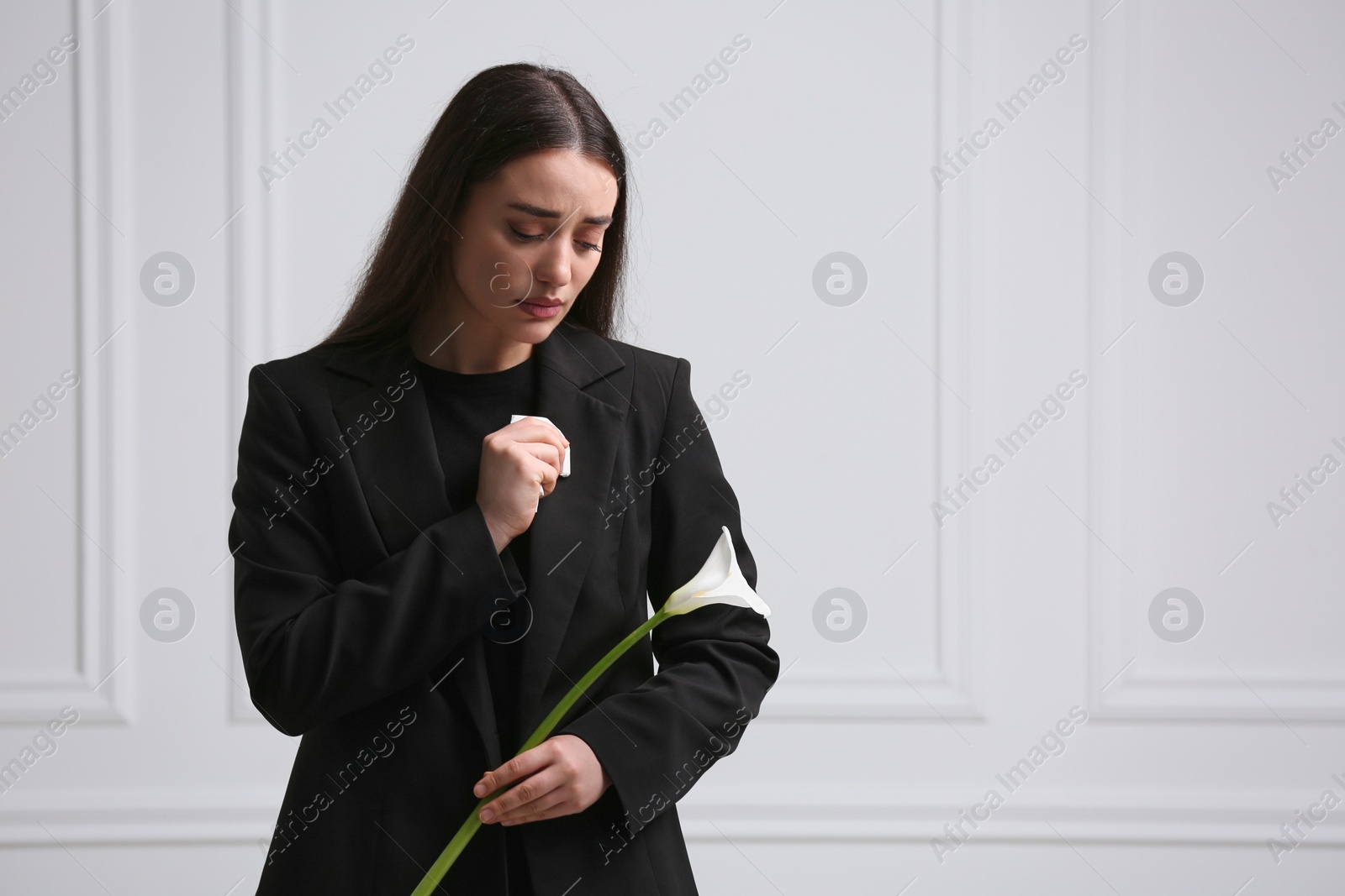 Photo of Sad woman with calla lily flower near white wall, space for text. Funeral ceremony