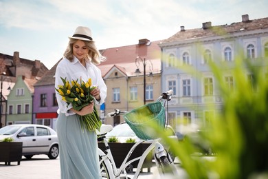 Beautiful woman with bouquet of yellow tulips and bicycle on city street