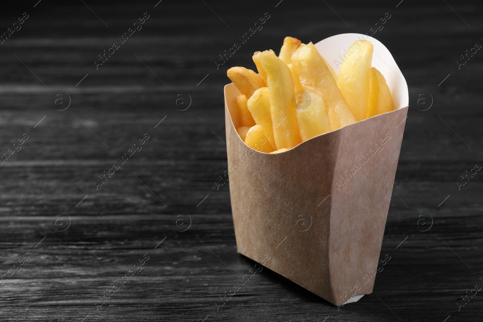 Photo of Delicious french fries in paper box on black wooden table, closeup. Space for text