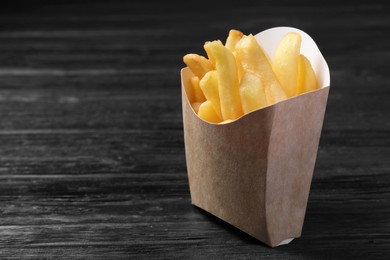 Delicious french fries in paper box on black wooden table, closeup. Space for text