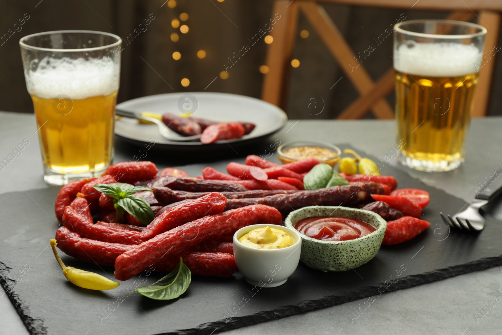 Photo of Different thin dry smoked sausages, sauces and glasses of beer on grey table