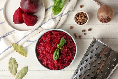 Photo of Bowl of grated boiled beets with basil, bay leaves and allspice on white wooden table, flat lay