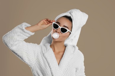 Photo of Young woman in bathrobe and sunglasses blowing chewing gum on brown background