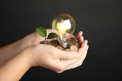 Photo of Woman with coins, light bulb and green plant against black background, closeup. Power saving