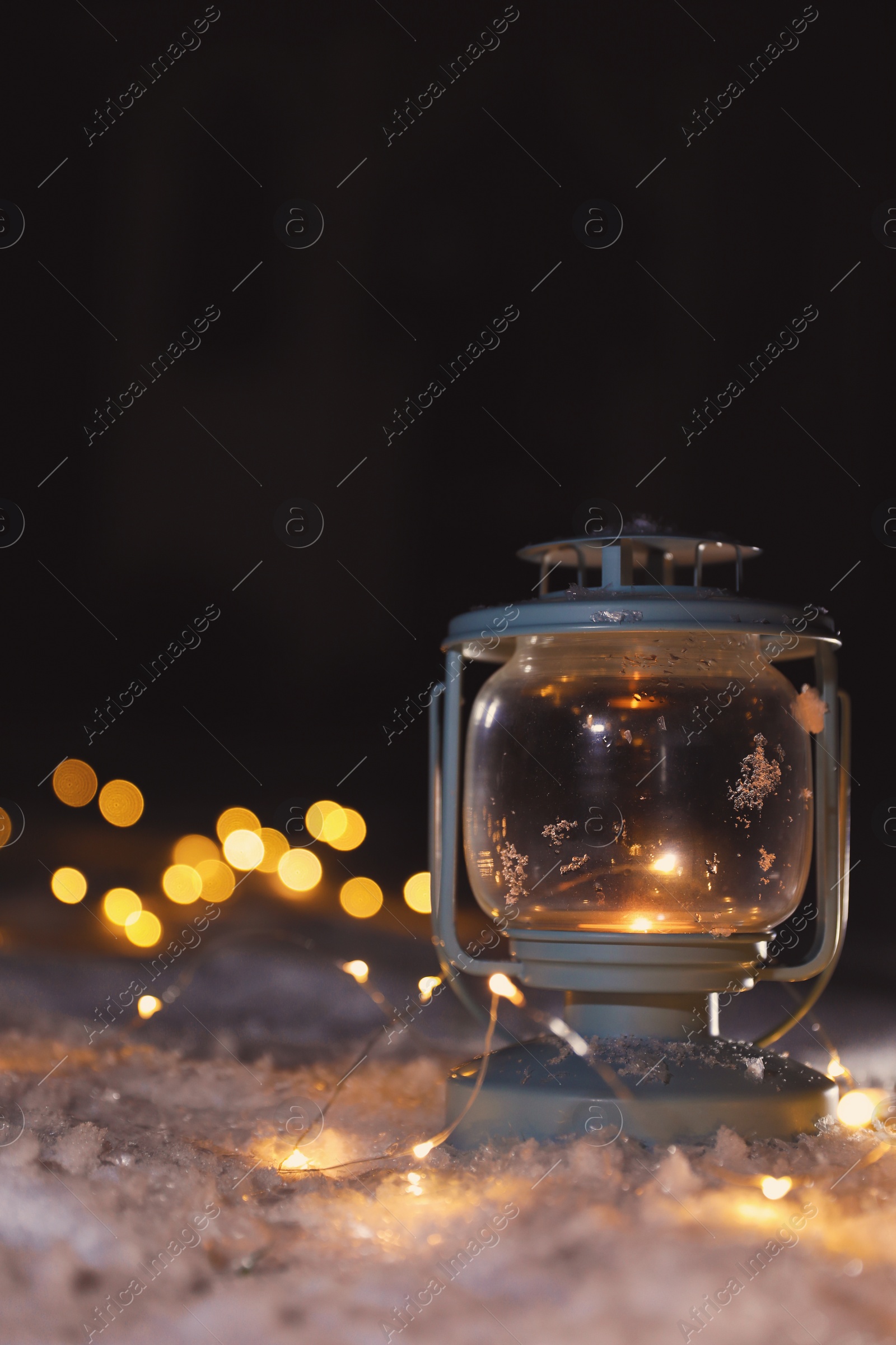 Photo of Lantern with burning candle and Christmas lights on white snow outdoors