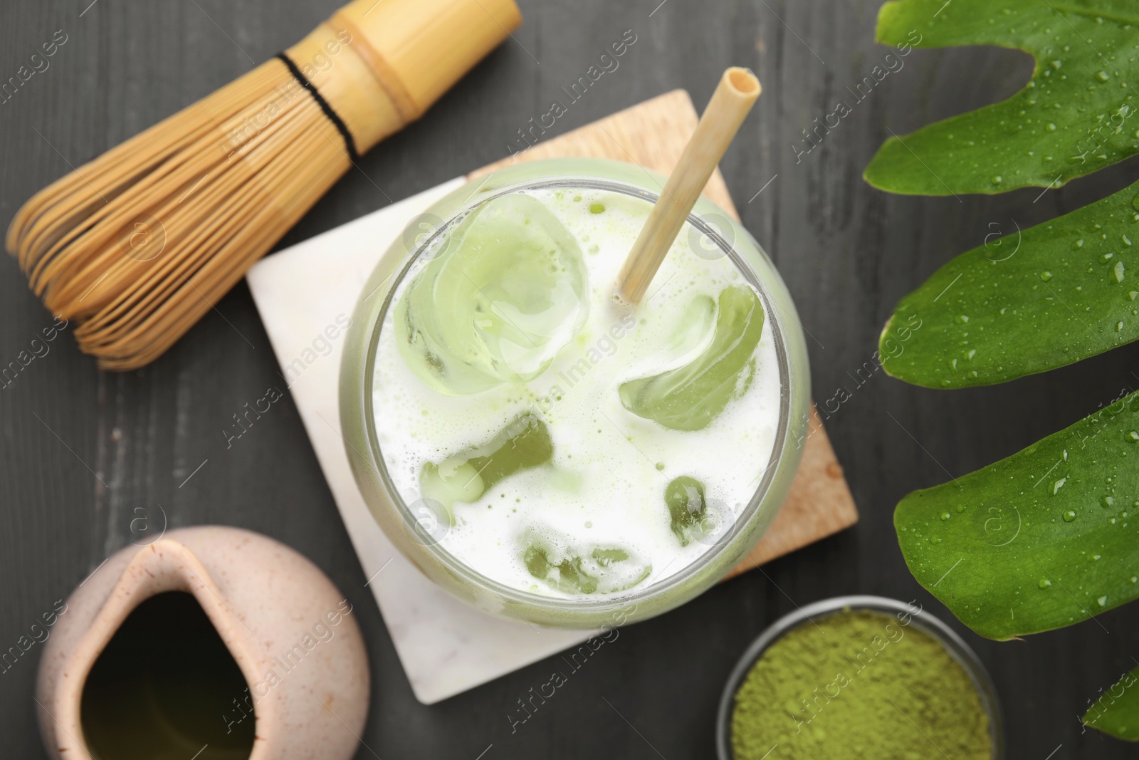 Photo of Glass of tasty iced matcha latte, bamboo whisk, leaf and powder on grey wooden table, flat lay