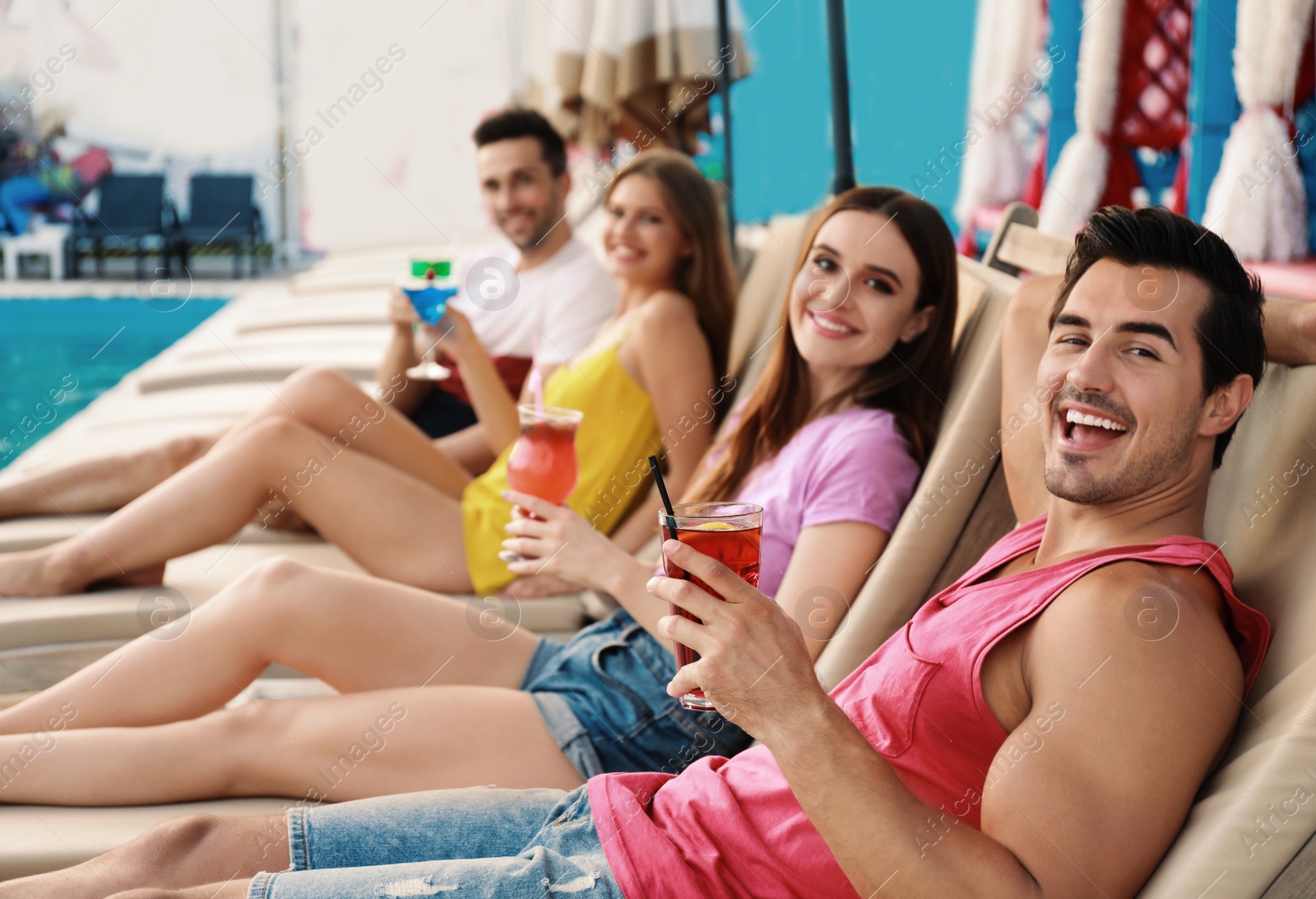 Photo of Happy young friends with fresh summer cocktails relaxing on sunbeds near swimming pool