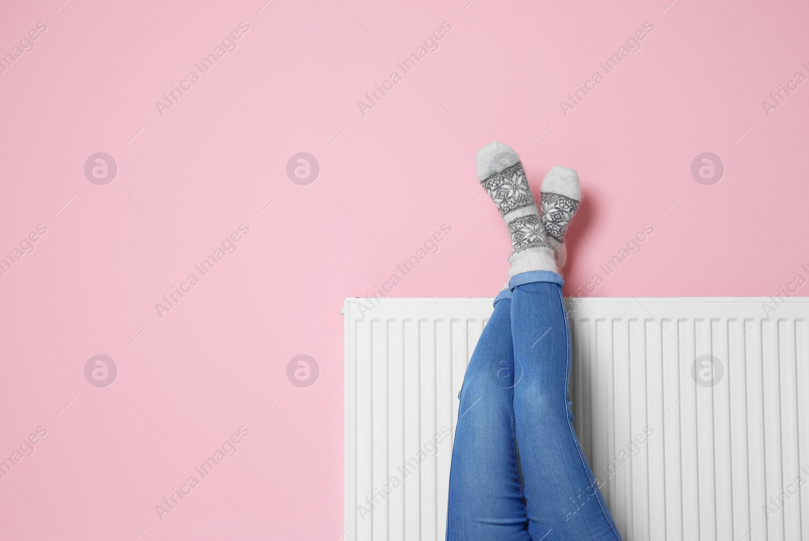Photo of Woman warming legs on heating radiator near color wall. Space for text