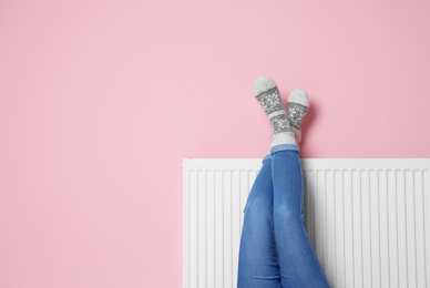 Woman warming legs on heating radiator near color wall. Space for text