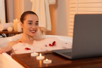 Photo of Happy woman with laptop taking bath in tub with foam and rose petals indoors