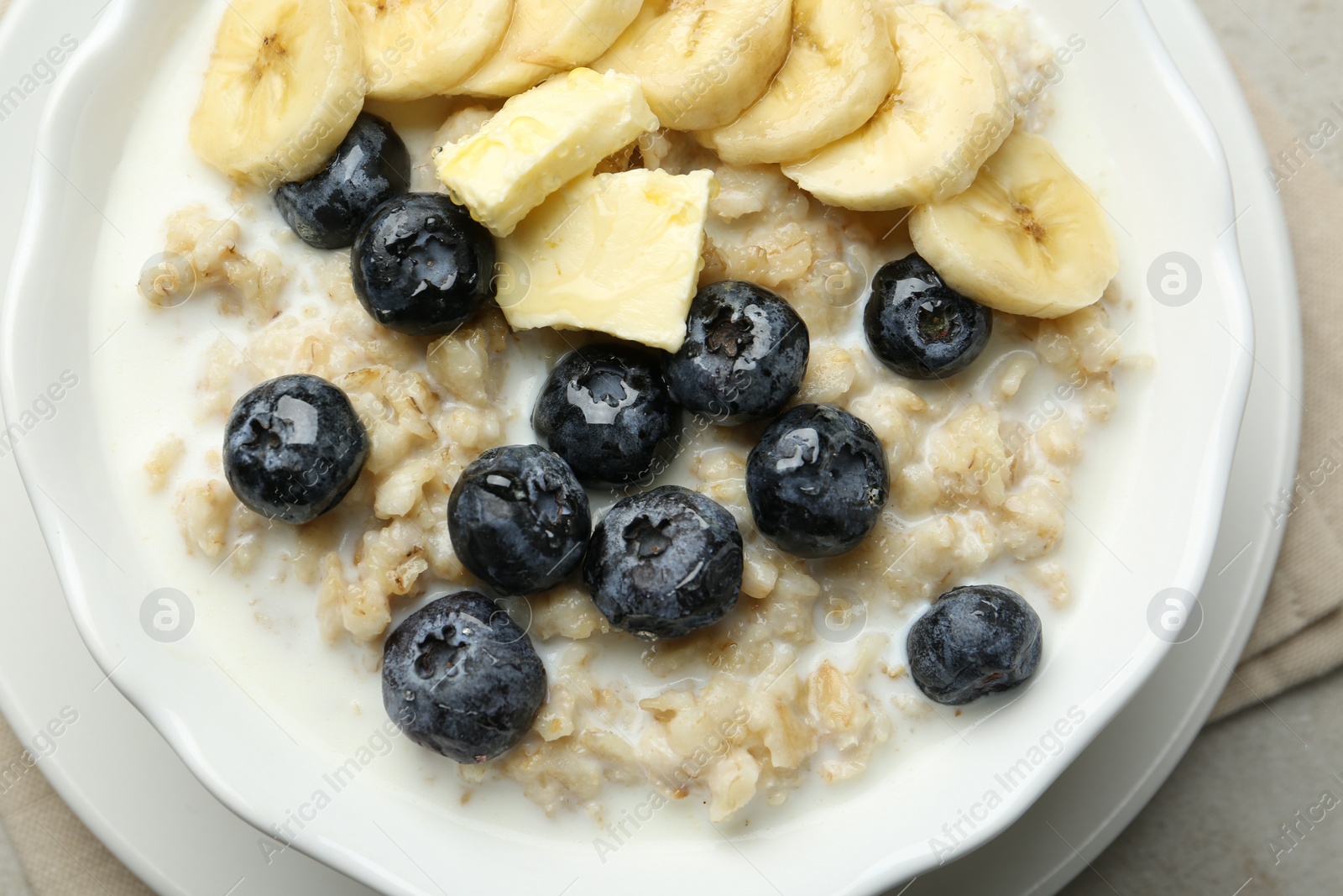 Photo of Tasty oatmeal with banana, blueberries, butter and milk served in bowl on light grey table, top view