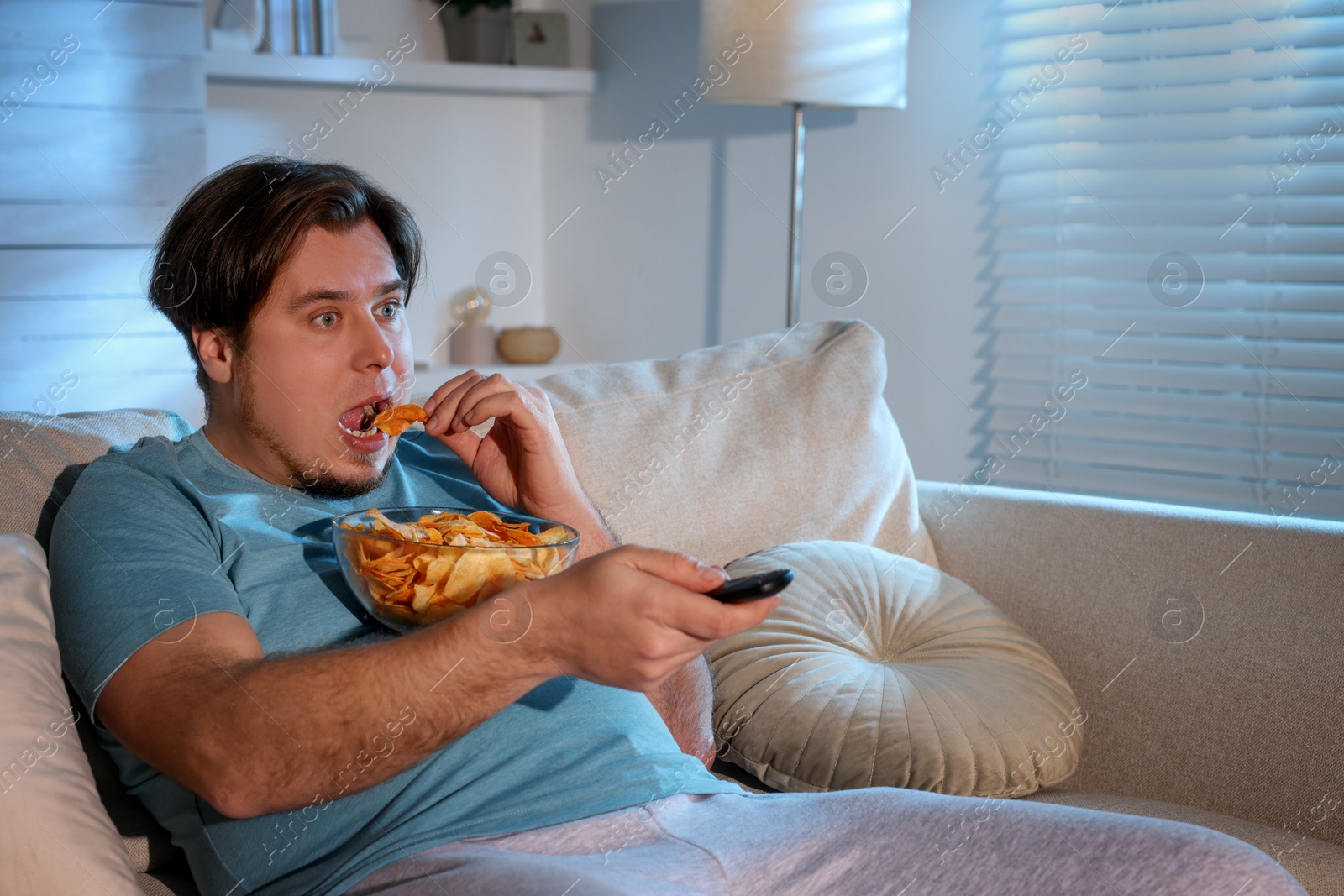 Photo of Overweight man with chips watching TV on sofa at home