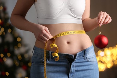 Woman with Christmas ball and measuring tape indoors, closeup. Overweight problem after New Year party