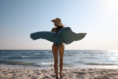 Photo of Woman with beach towel and straw hat on sand near sea, back view