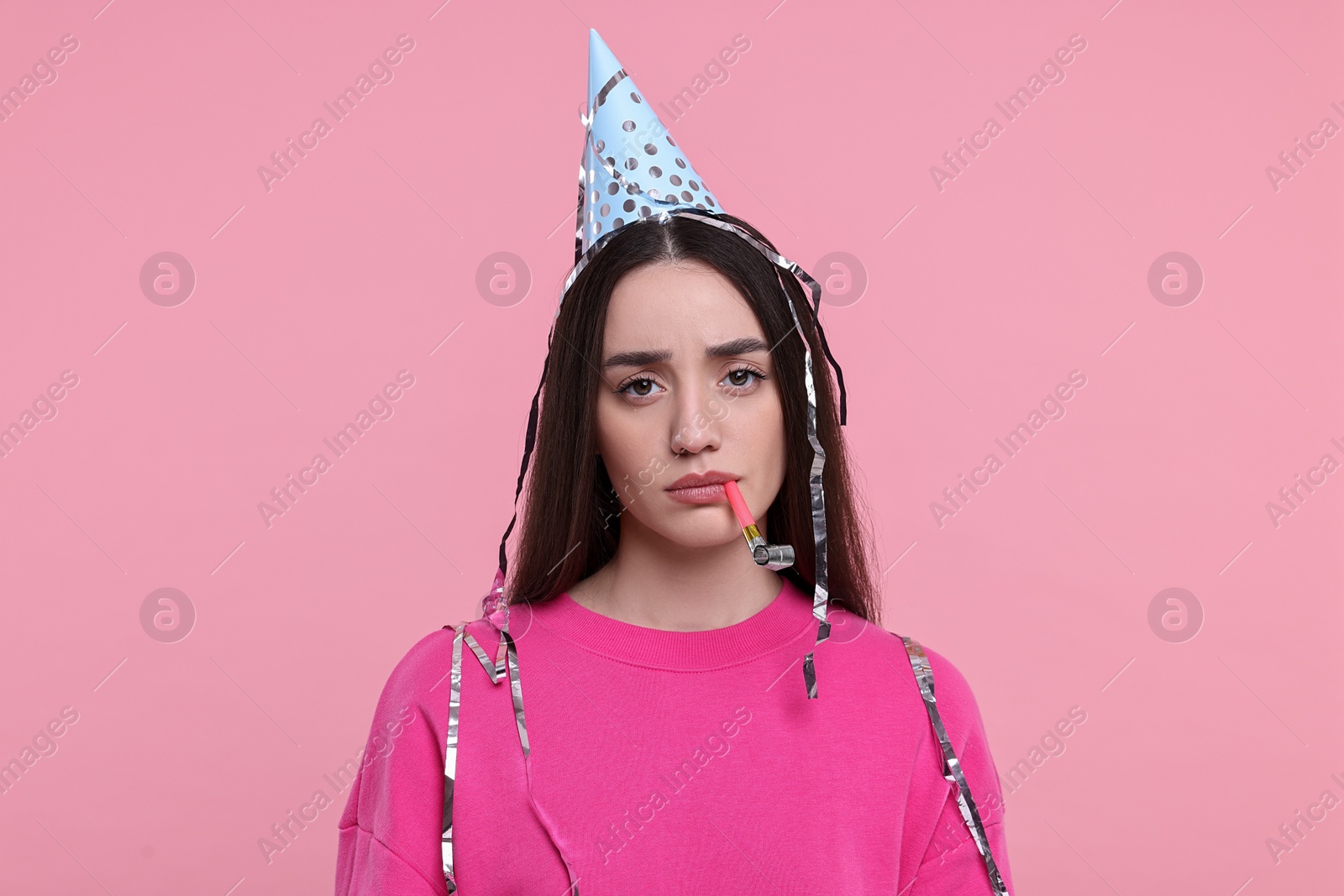 Photo of Sad woman in party hat with blower and streamers on pink background
