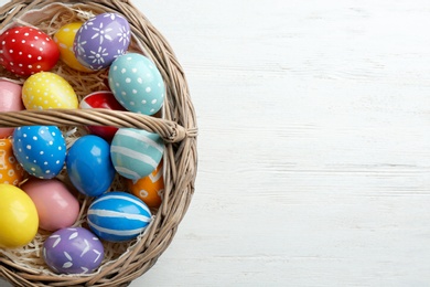Photo of Wicker basket with painted Easter eggs on wooden table, top view. Space for text