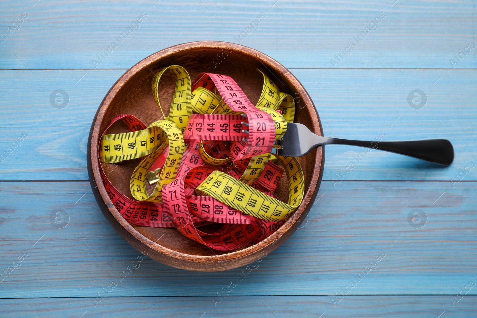 Photo of Bowl with measuring tape and fork on light blue wooden table, top view. Weight loss concept