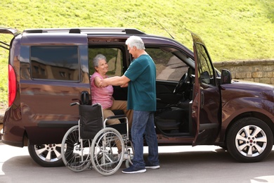 Photo of Mature man helping senior woman to get out from van into wheelchair outdoors