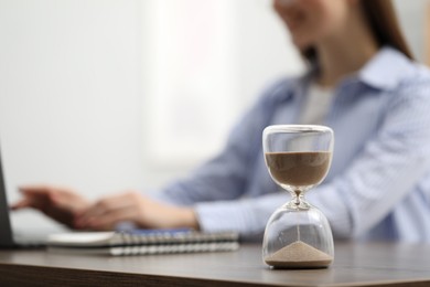 Photo of Hourglass with flowing sand on desk. Woman using laptop indoors, selective focus