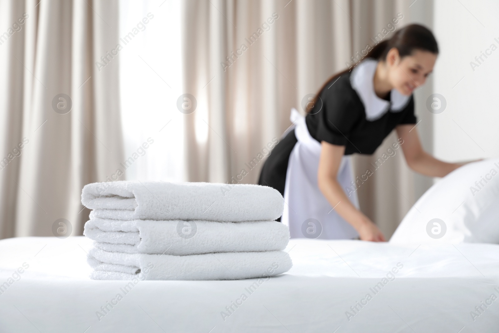 Photo of Young maid making bed in hotel room, focus on stack of towels