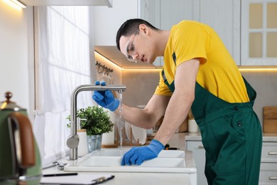 Photo of Young plumber wearing protective glasses and gloves examining faucet in kitchen