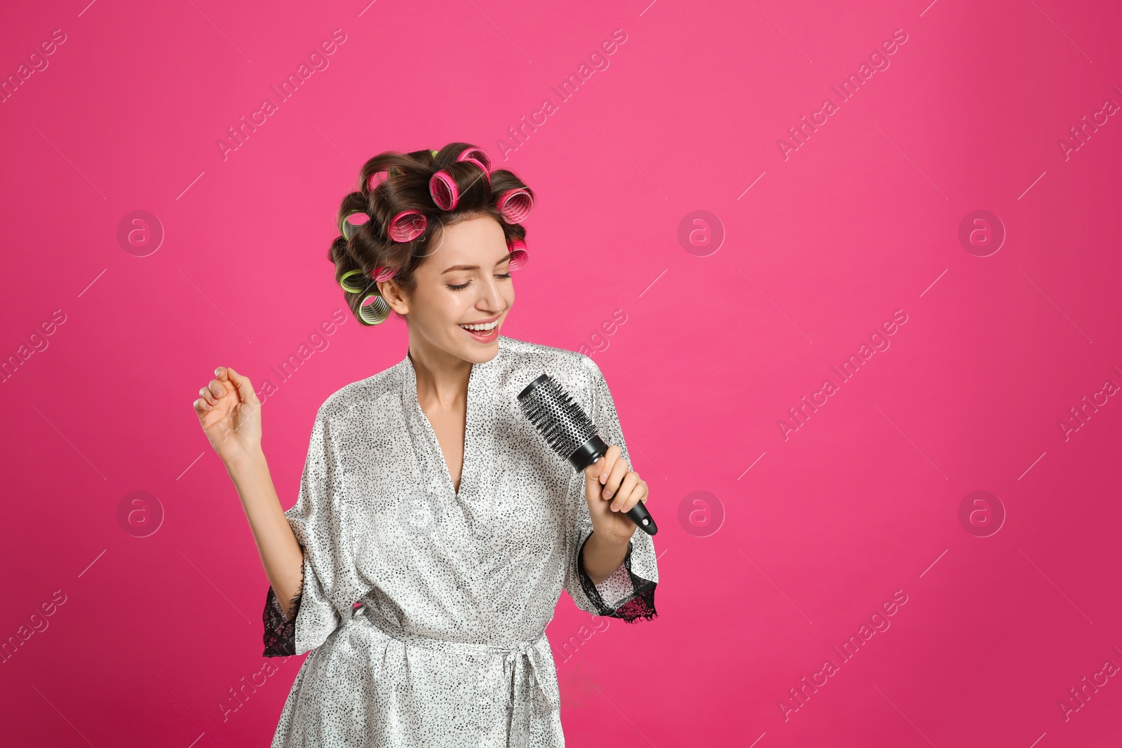 Photo of Beautiful young woman in silk bathrobe with hair curlers singing into hairbrush on pink background. Space for text