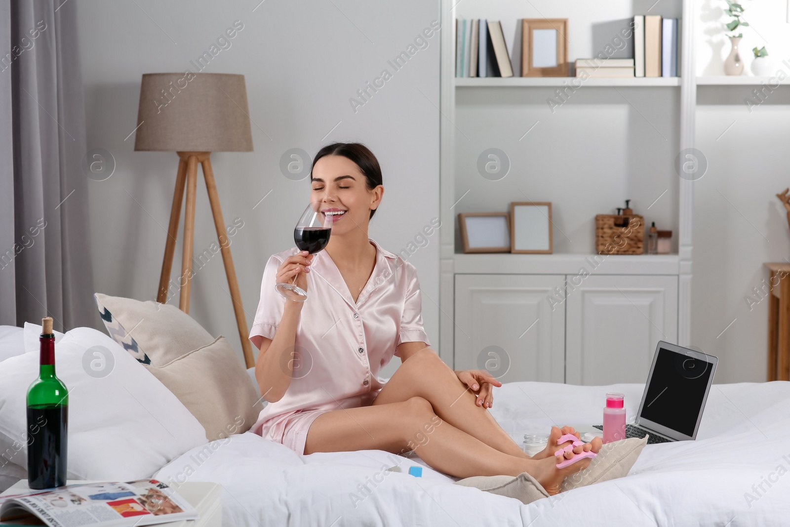 Photo of Beautiful young woman with glass of wine giving herself pedicure on bed at home