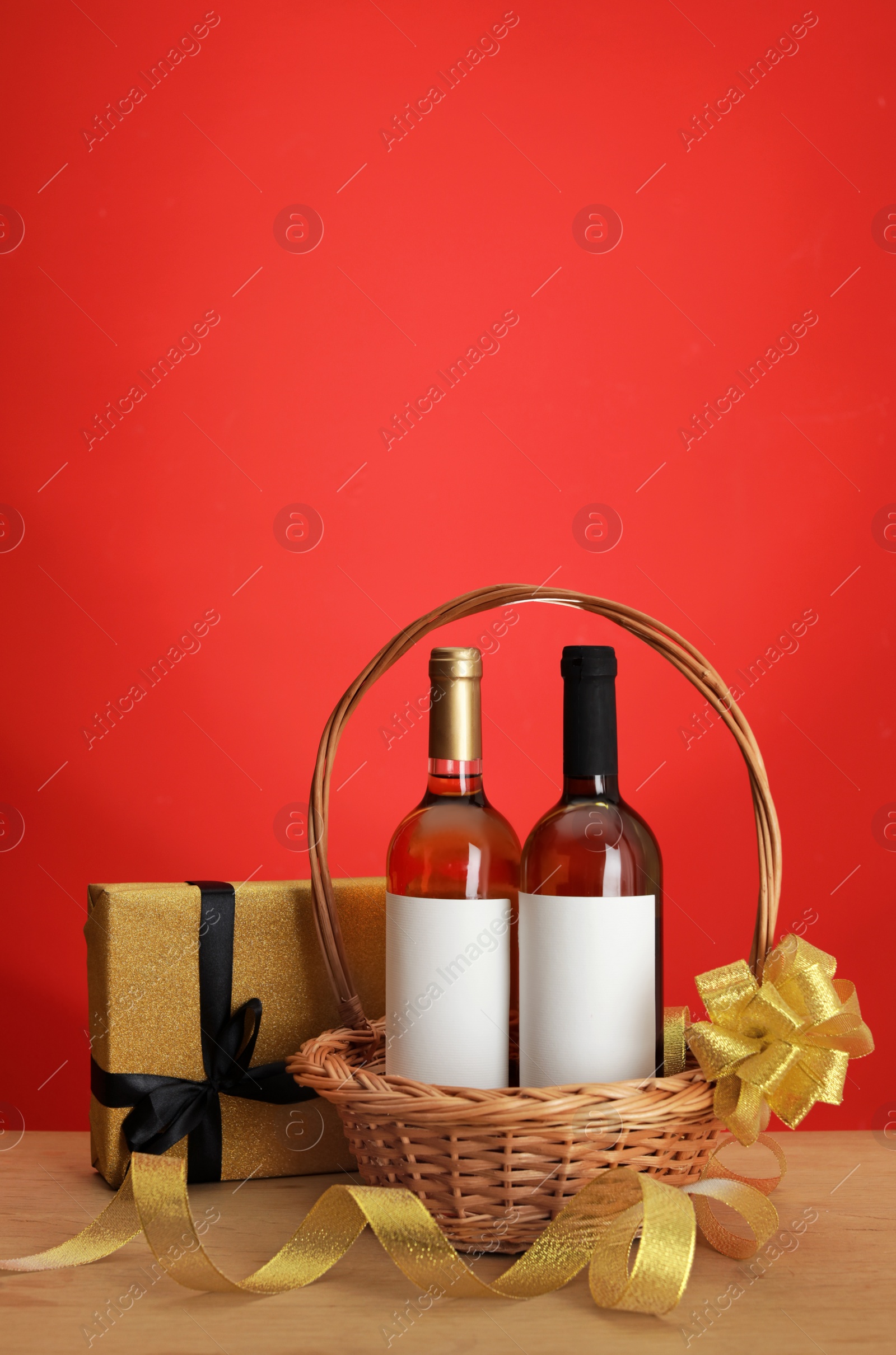 Photo of Bottles of wine in wicker basket with bow and gift on table against color background. Space for text