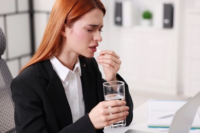 Woman with pill and glass of water suffering from headache at table in office, space for text