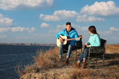 Couple with guitar resting in camping chairs near river on sunny day