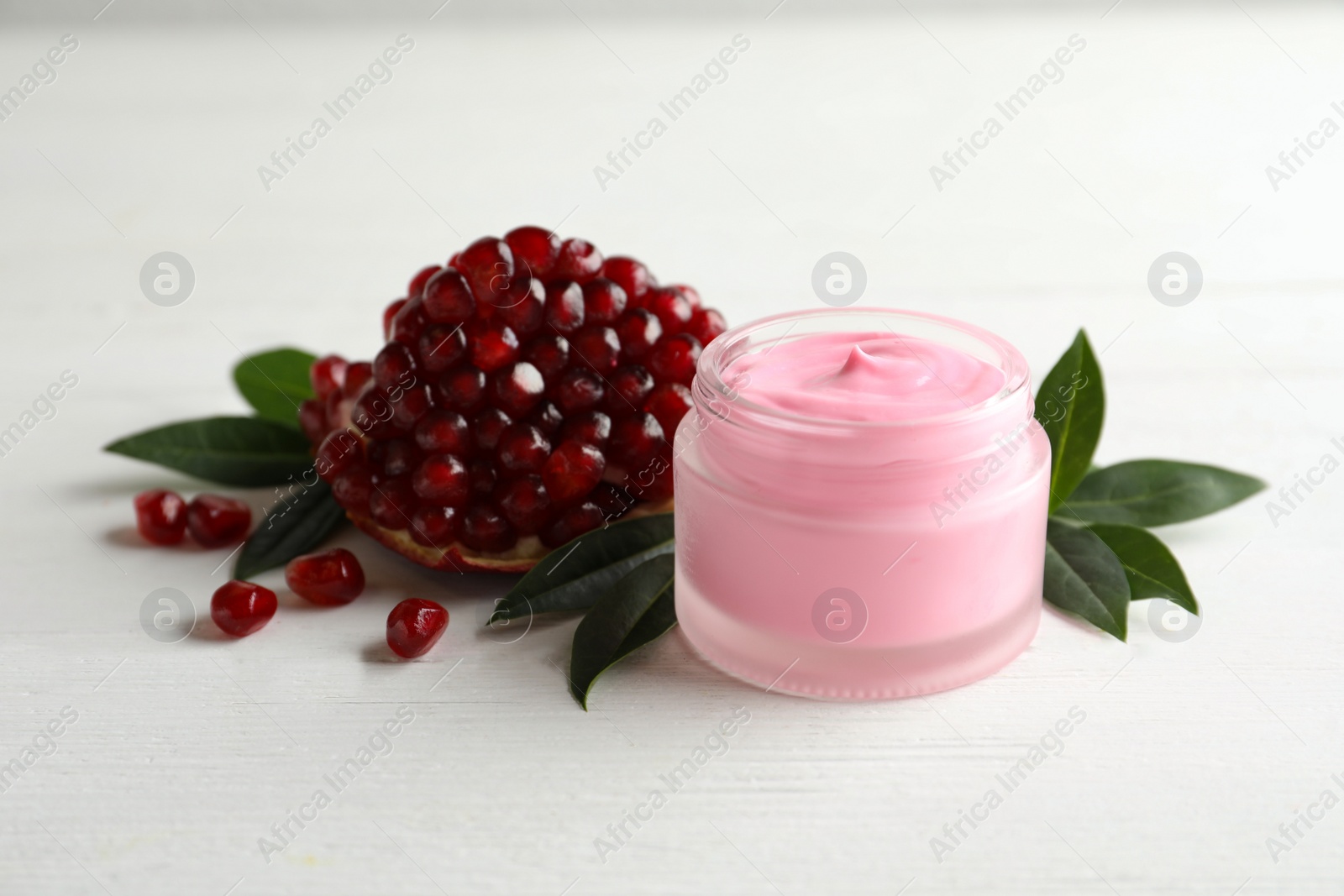 Photo of Jar with natural facial mask, pomegranate seeds and green leaves on white wooden table, closeup
