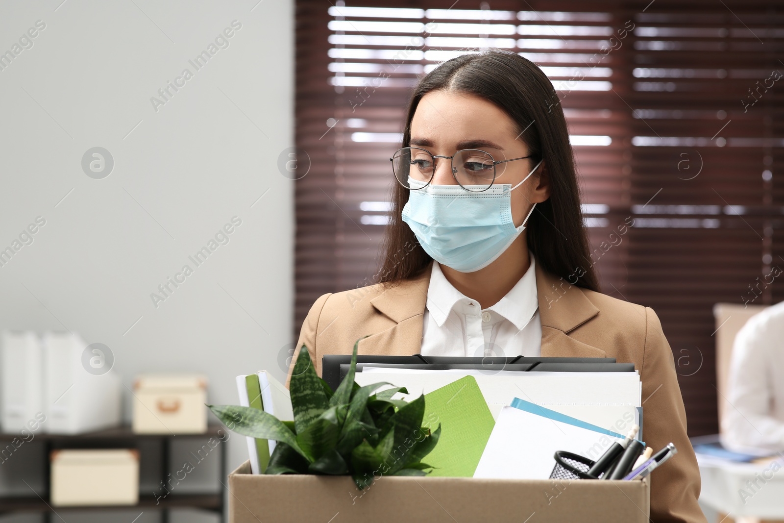 Photo of Upset dismissed woman wearing protective mask carrying box with personal stuff in office