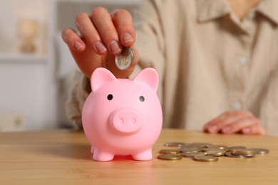 Photo of Woman putting coin into pink piggy bank at wooden table, closeup