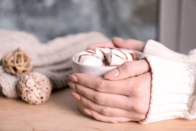 Photo of Woman in sweater holding cup of aromatic cacao with marshmallows on wooden table, closeup. Space for text
