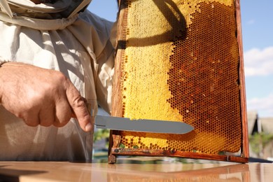 Photo of Senior beekeeper uncapping honeycomb frame with knife at table outdoors, closeup