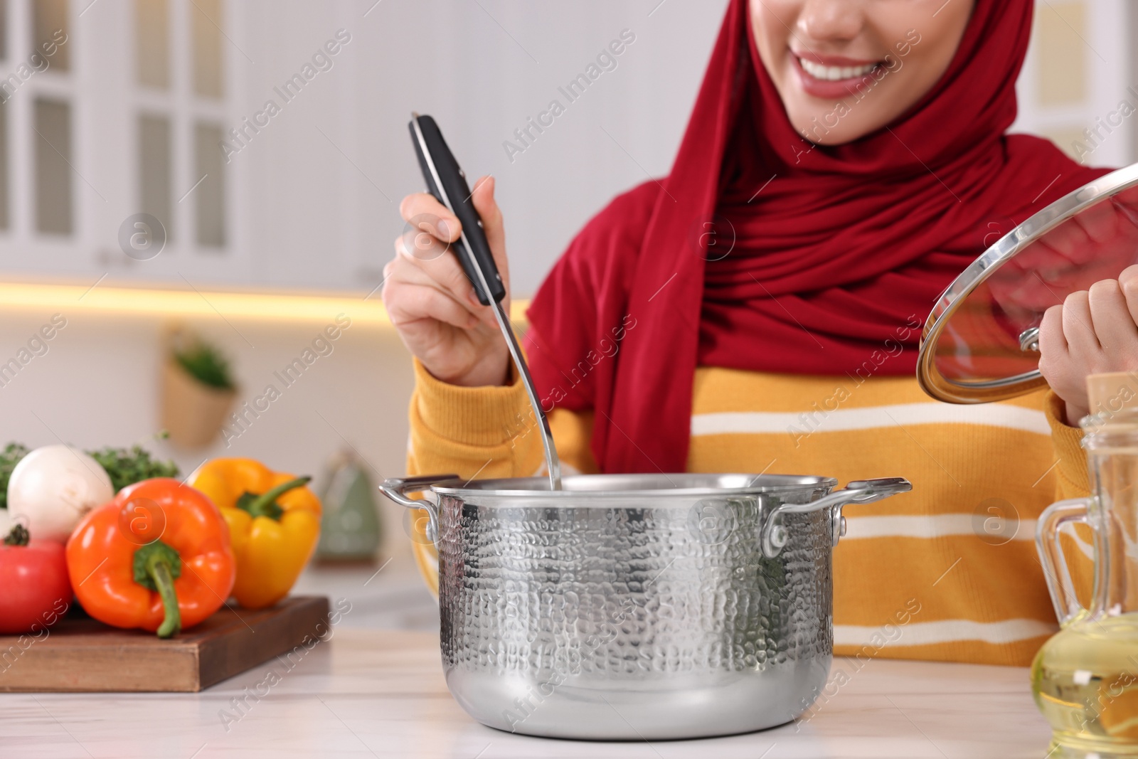 Photo of Muslim woman making delicious soup with vegetables at white table in kitchen, closeup