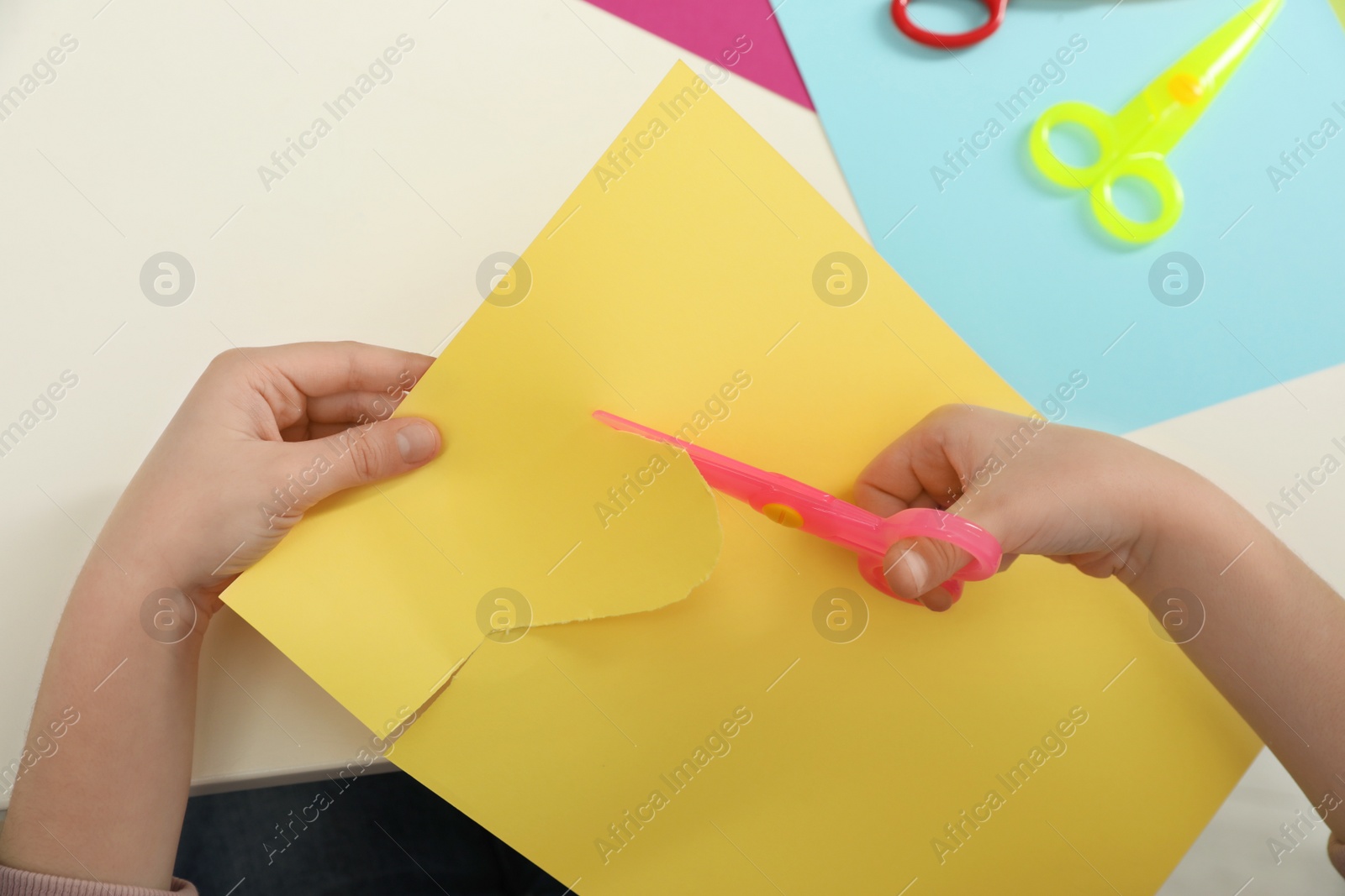 Photo of Child cutting color paper with plastic scissors at table, closeup