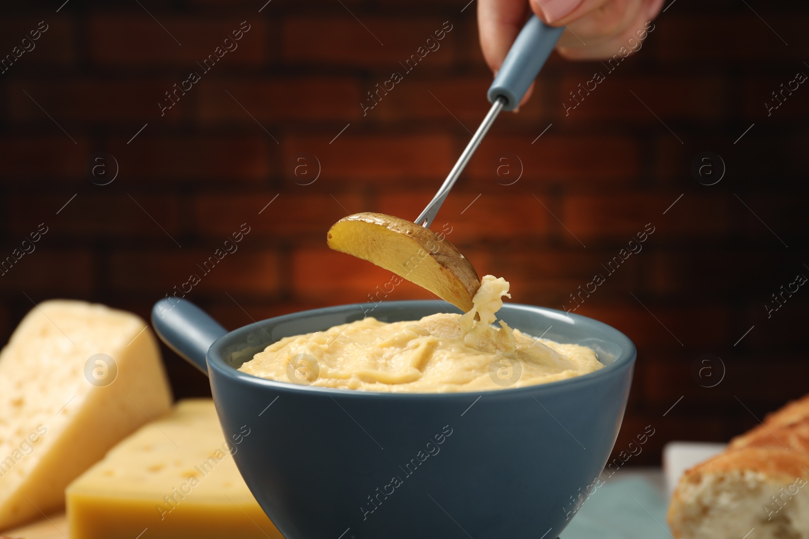 Photo of Woman dipping piece of potato into fondue pot with melted cheese on blurred background, closeup