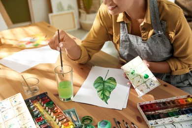 Photo of Young woman drawing leaf with watercolors at table indoors, closeup