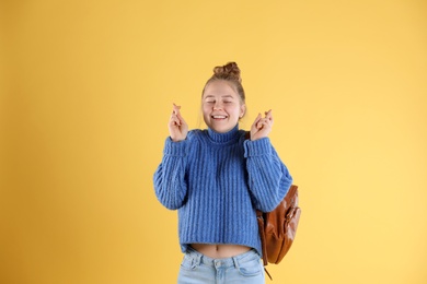 Pretty teenage girl with backpack crossing her fingers on color background