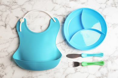 Set of colorful tableware and silicone bib on white marble background, flat lay. Serving baby food